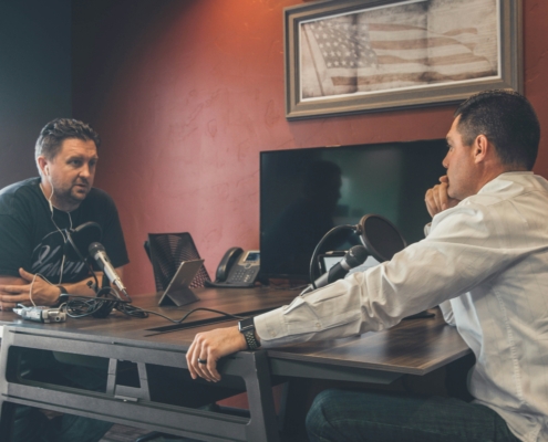 Two men recording a podcast at a table, one speaking into a microphone, the other listening attentively.