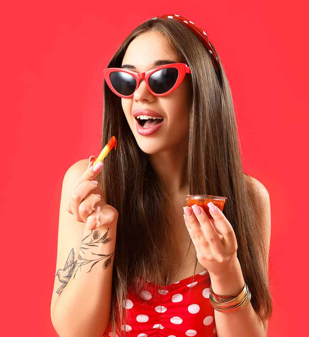 young woman eating french fries with ketchup on red background, wearing sunglasses and sundress