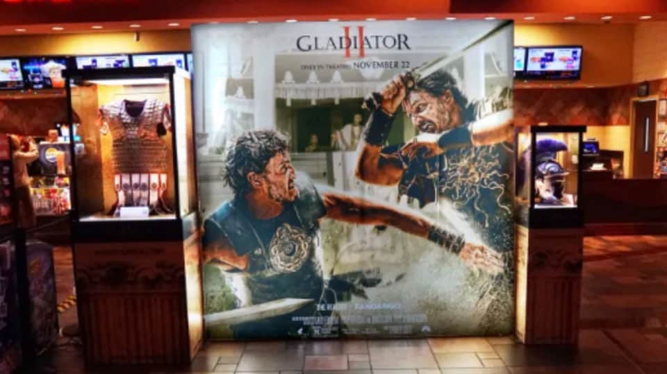 gladiator II movie poster in theater, with movie props on either side in glass cases, for pr campaign for film’s release